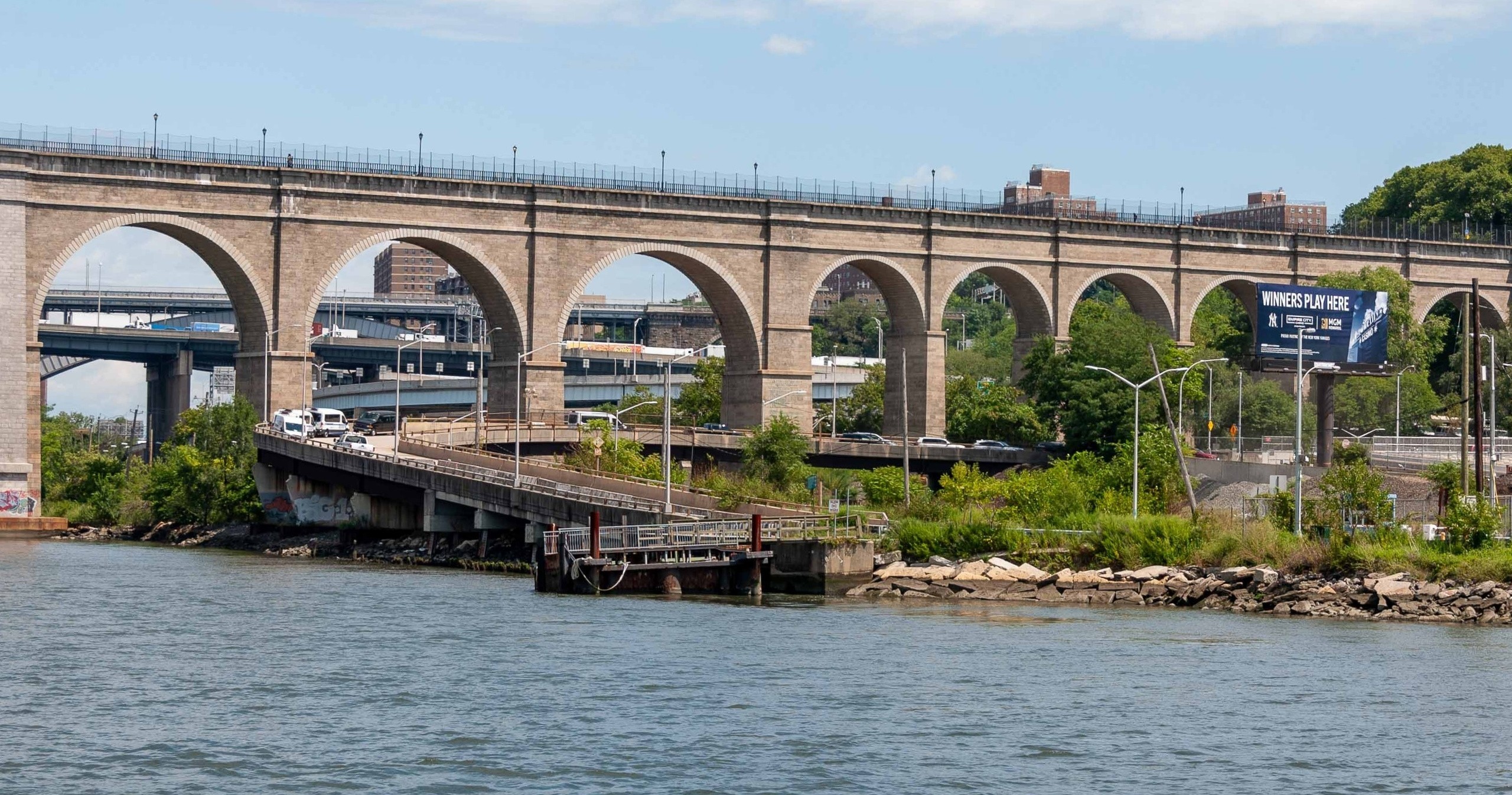 Stone arched bridge over river with highway and buildings in background.