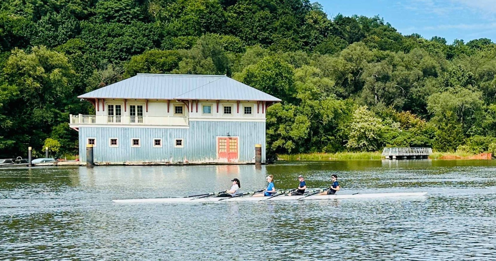 Four people rowing near a blue boathouse by a forest.