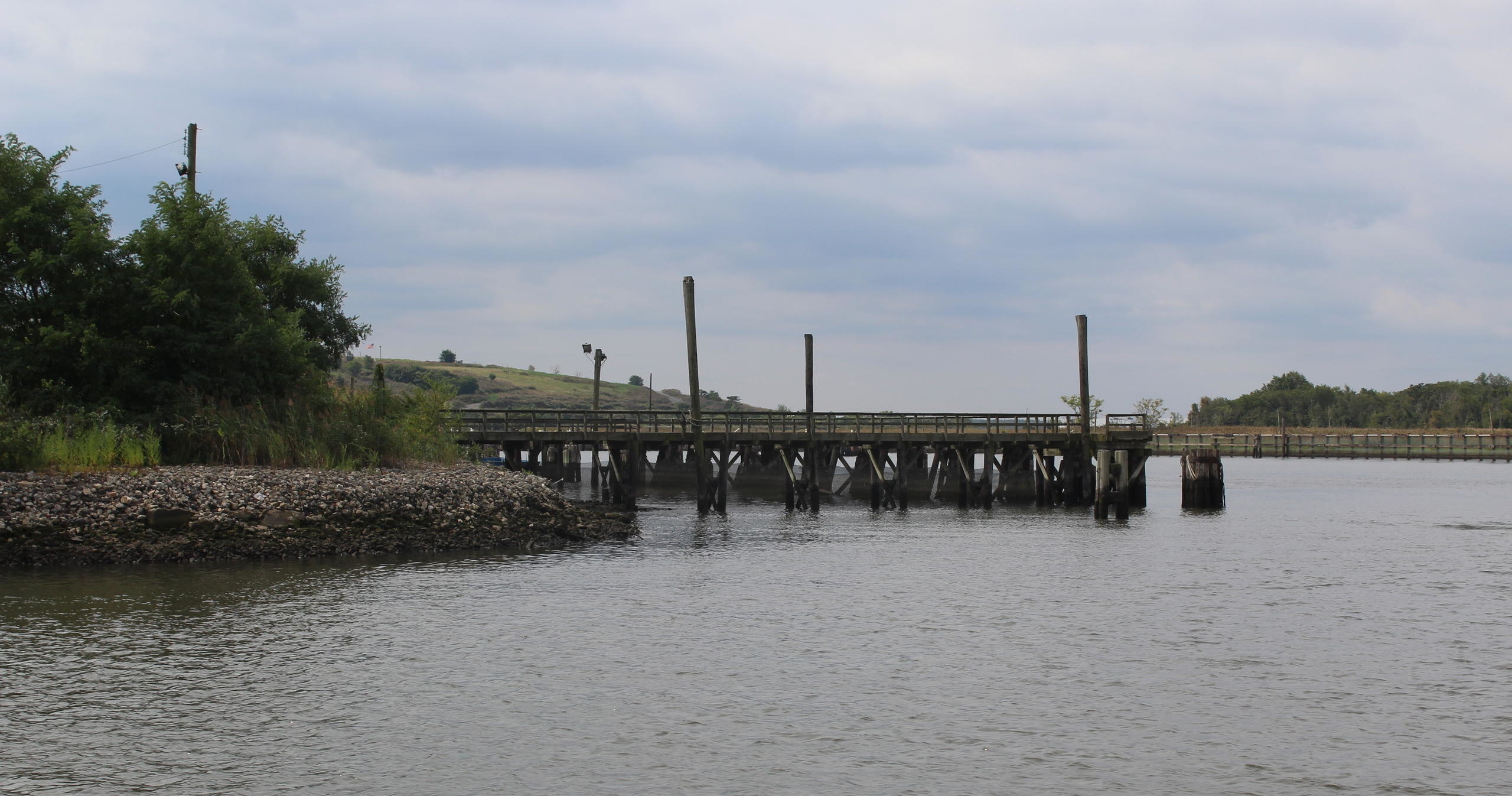 Wooden pier extending over water with cloudy sky and greenery nearby.