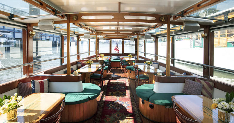 Interior of a boat with wooden tables, cushioned seating, and large windows.