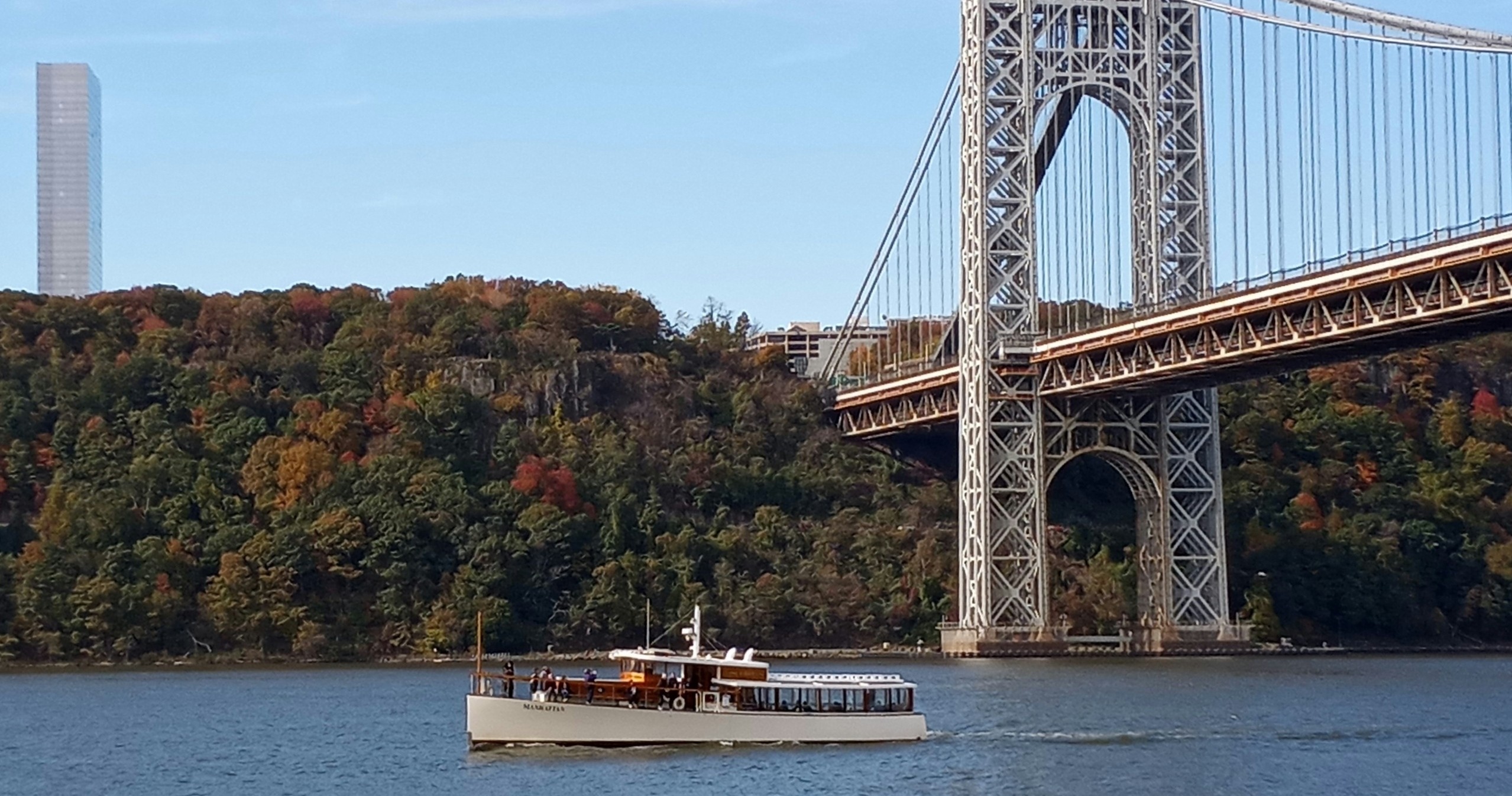 Boat on river near large suspension bridge and forested hill.