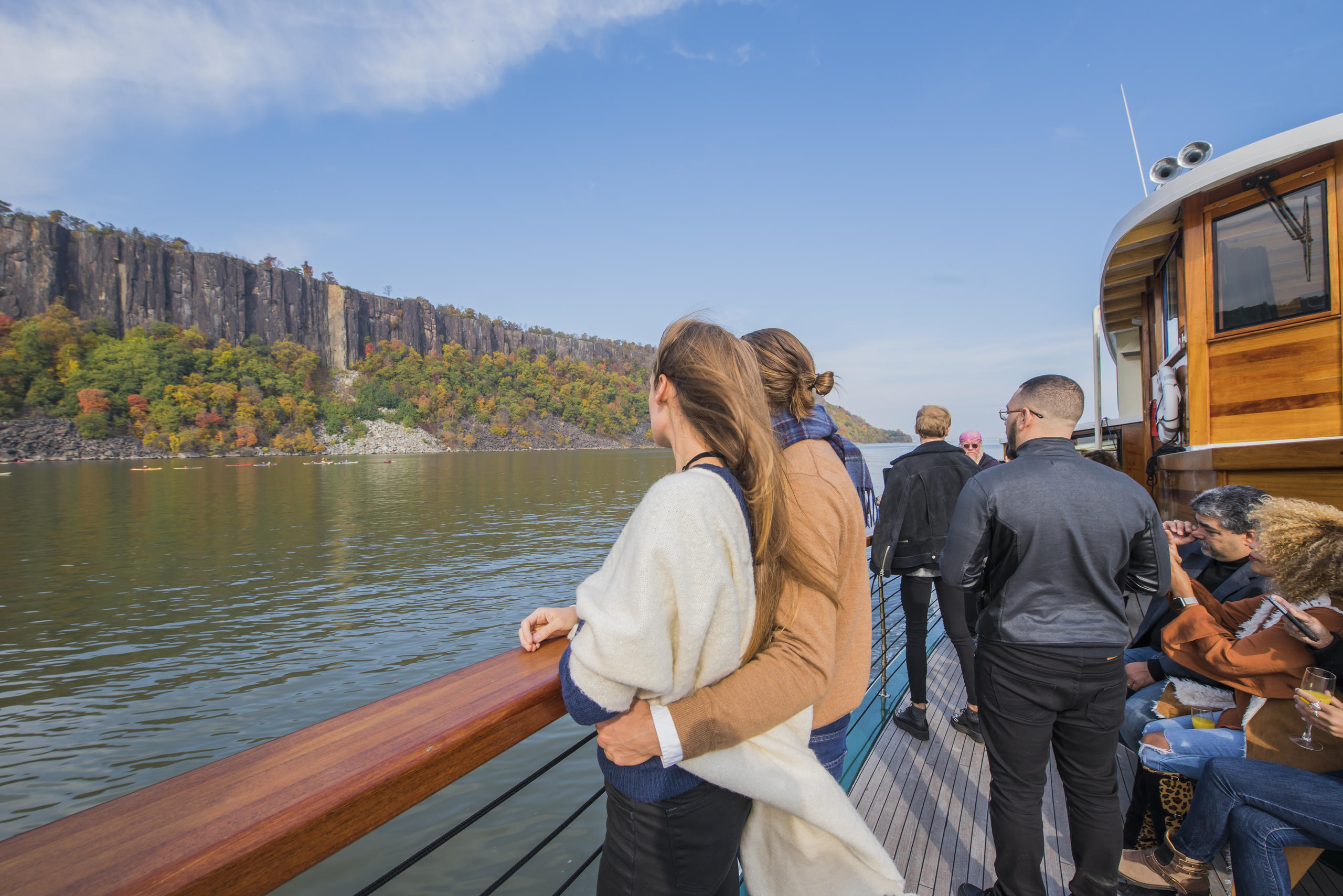 People enjoy river view from a boat, with scenic cliffs and autumn trees in the background.