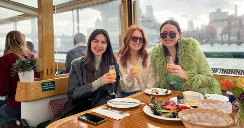 Three people smiling with drinks on a boat, table with food in front.