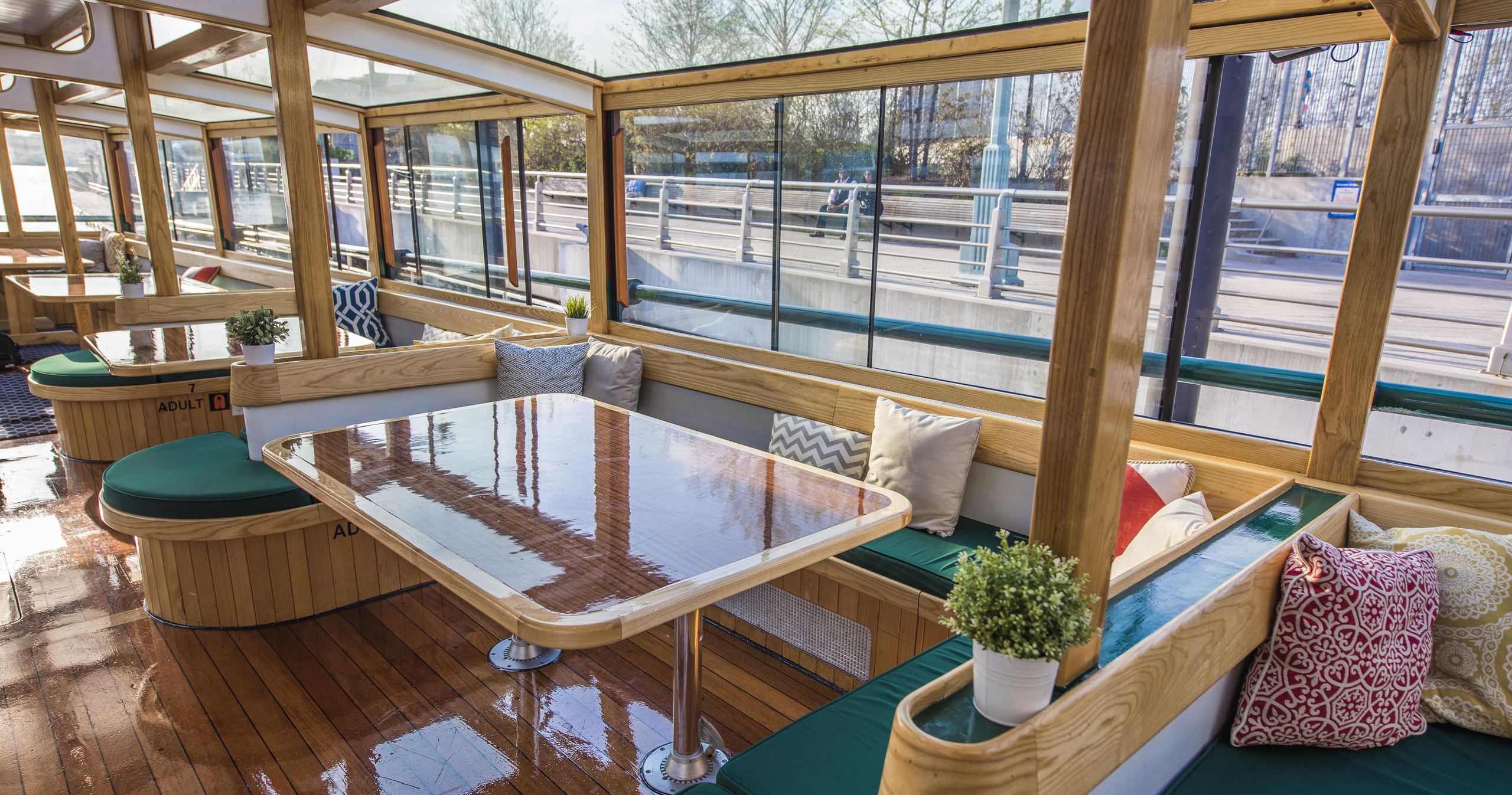 Boat interior with wooden tables, green-cushioned benches, and potted plants by large windows.