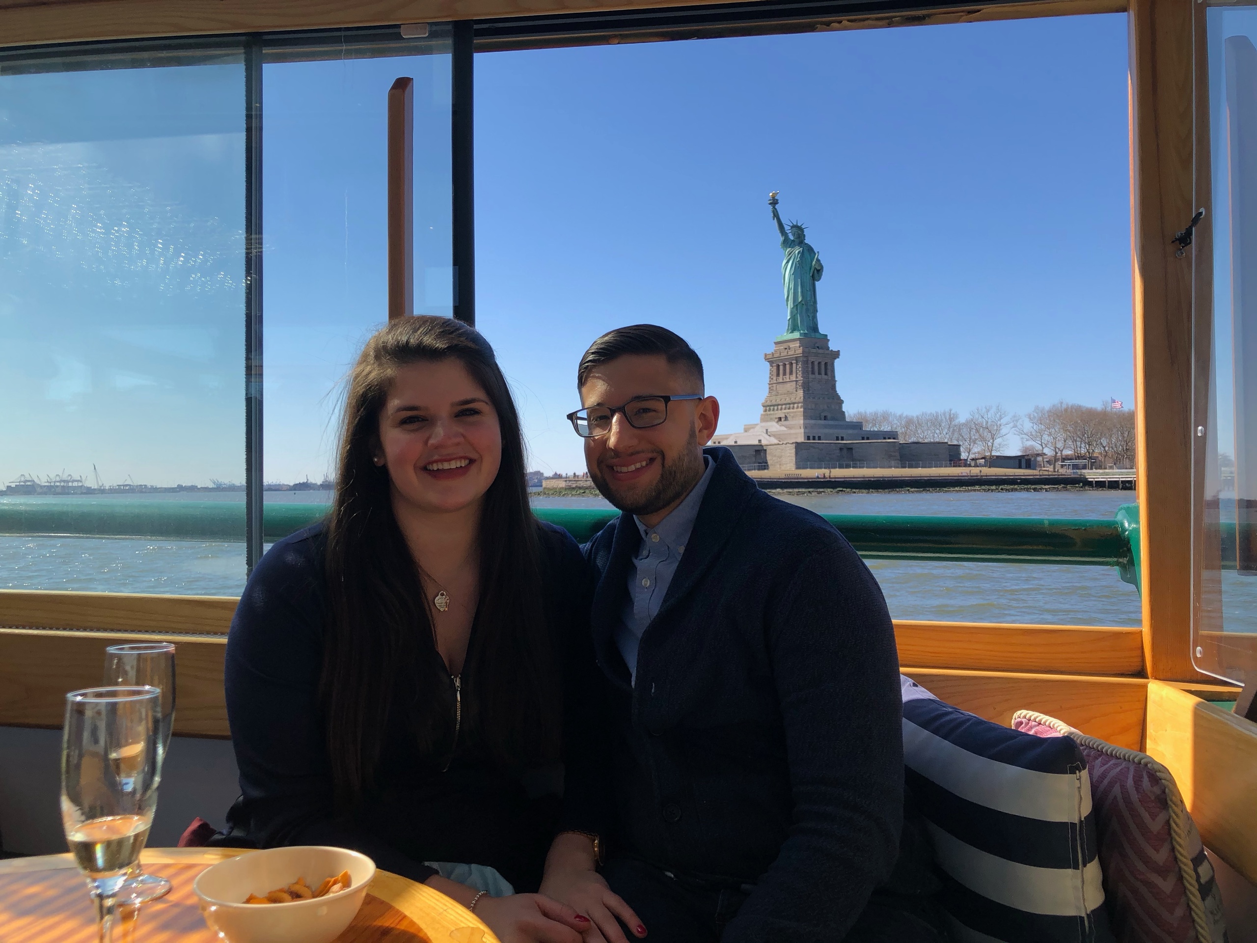 Couple smiling on a boat with the Statue of Liberty in the background.