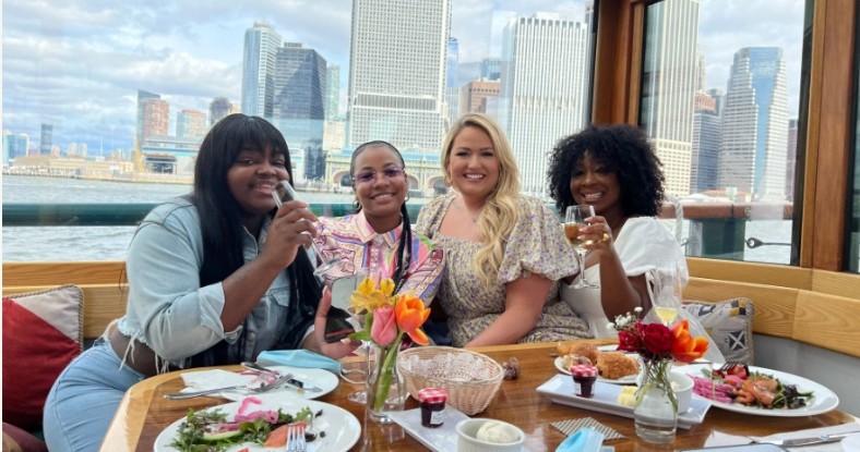 Four women smiling, holding drinks on a boat with city skyline in background.