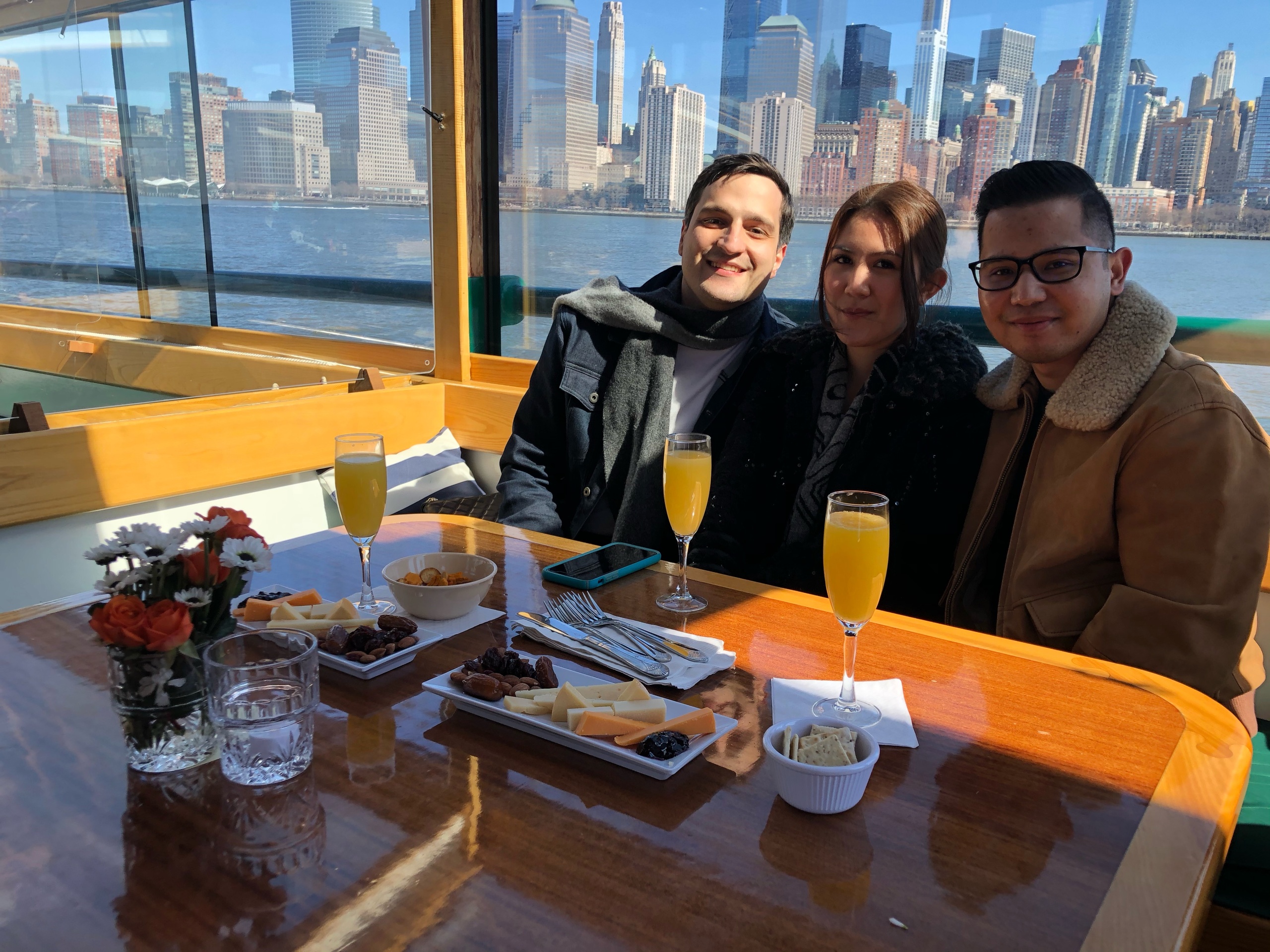 Three people smiling with drinks at a table, cityscape view in background.