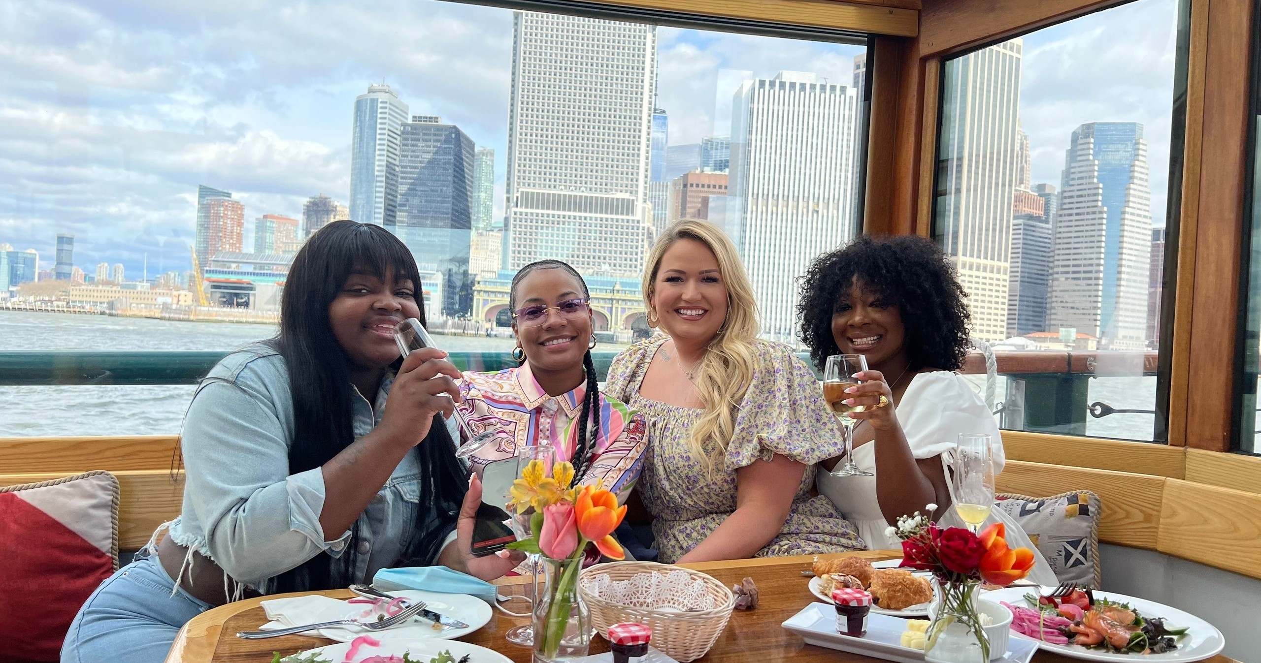 Four women smiling and holding drinks on a boat with city skyline in background.