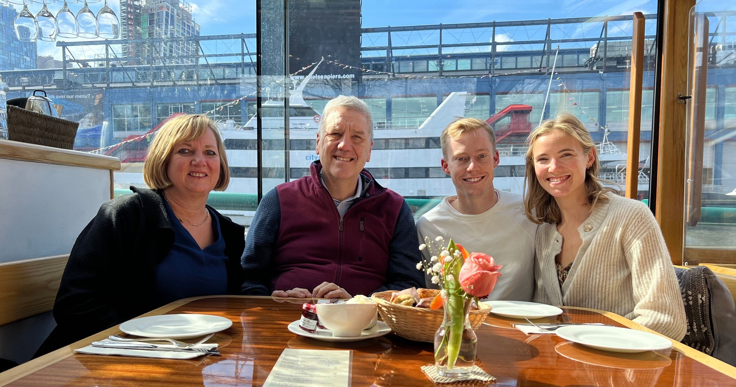 Four people seated at a dining table with a cityscape and boat in the background.