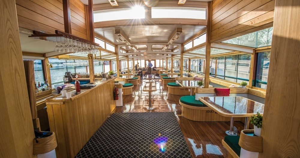 Interior of a wooden boat with tables, chairs, and large windows, sunlight streaming in.