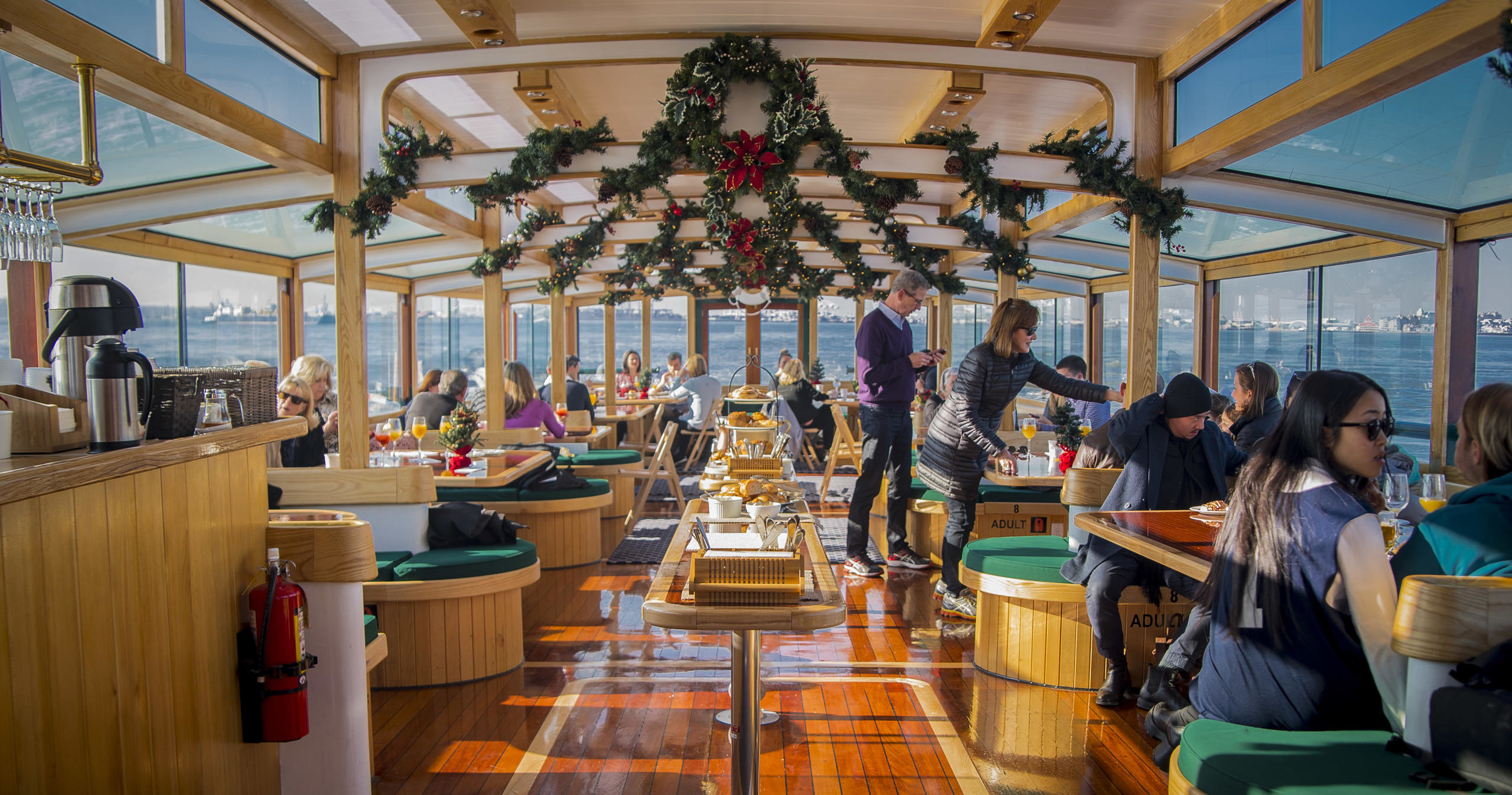 People dining inside a decorated boat with garlands and large windows overlooking the water.