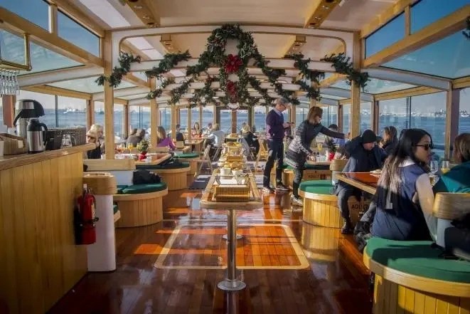 Decorated ferry interior with passengers seated, garlands hang above, windows show a water view.