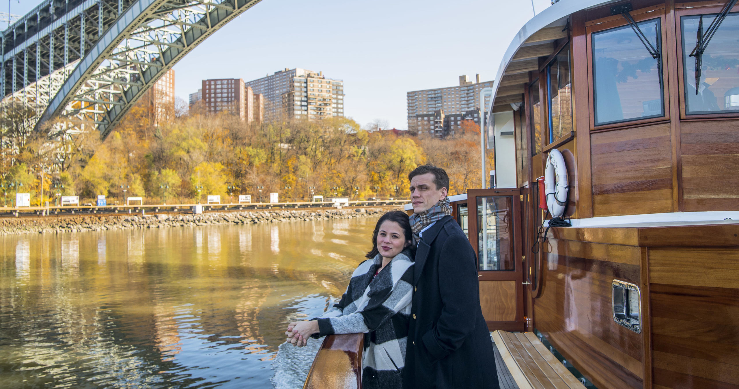 Two people on a boat under a bridge by autumn trees.
