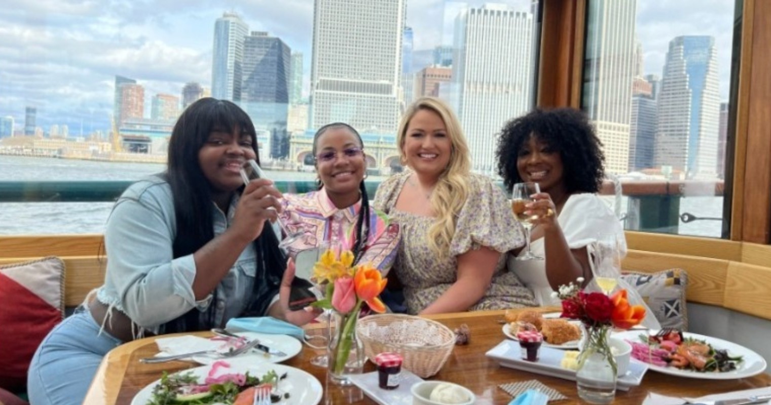 Four women smiling and holding drinks at a table with city skyline in background.
