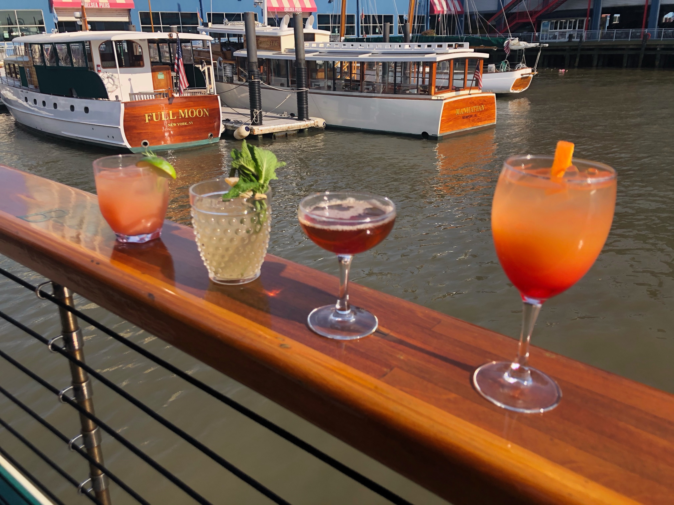 Four cocktails on a railing with boats on a river in the background.