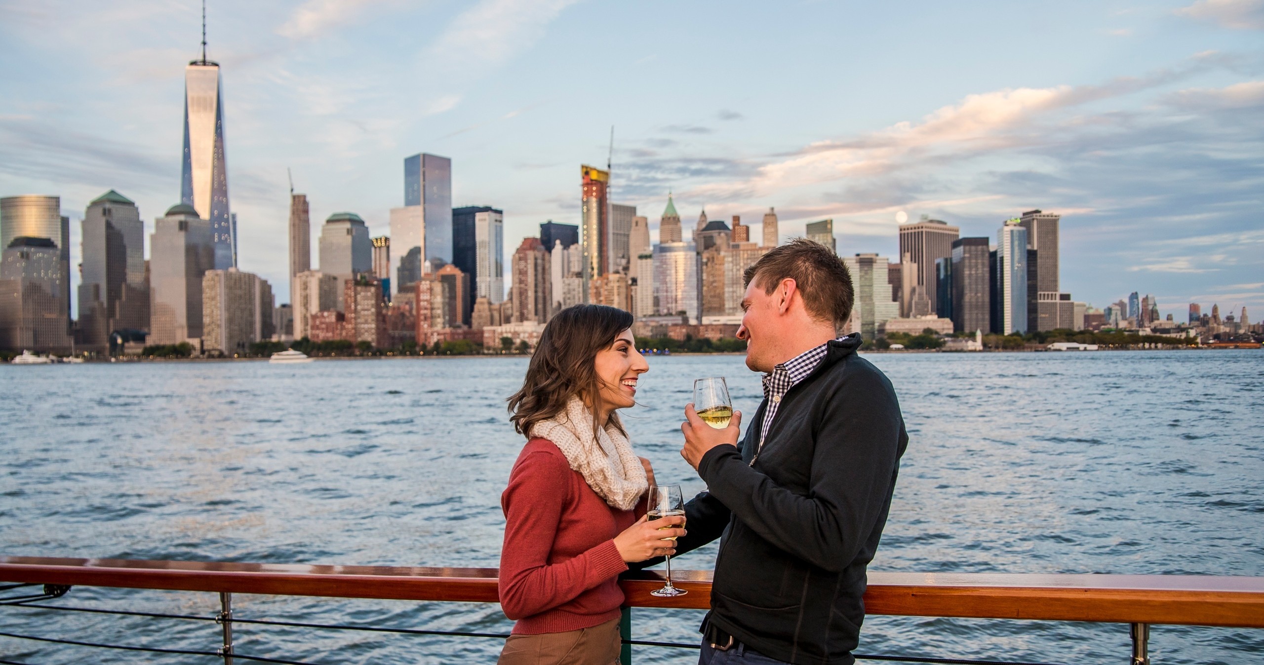 Couple on boat with wine glasses, NYC skyline in background.