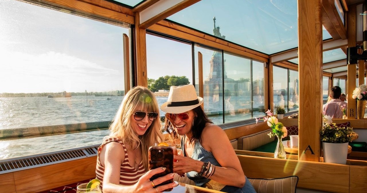 Two women taking a selfie on a boat with the Statue of Liberty in the background.