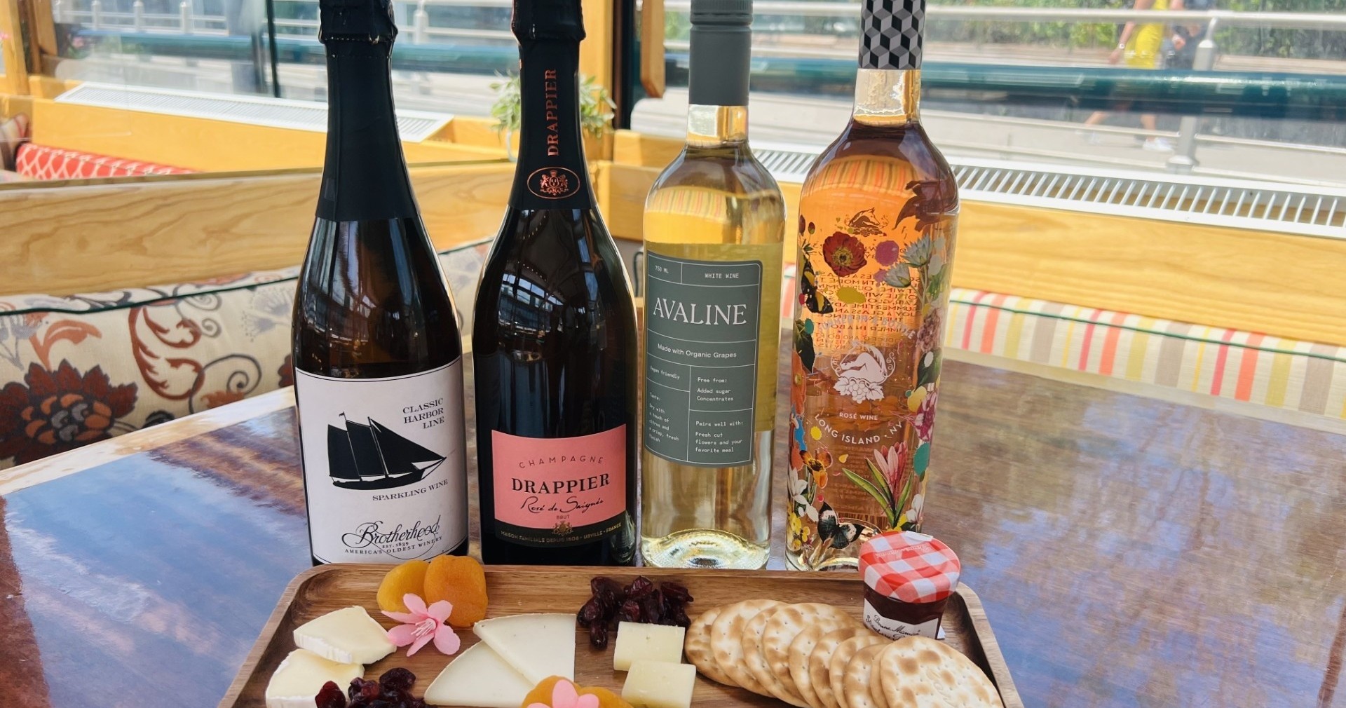 Four wine bottles and a tray with cheese, crackers, jam, and flowers on a table.