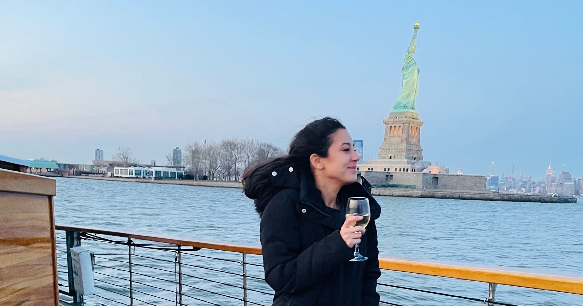 Person in a black coat holding a glass, with the Statue of Liberty in the background.