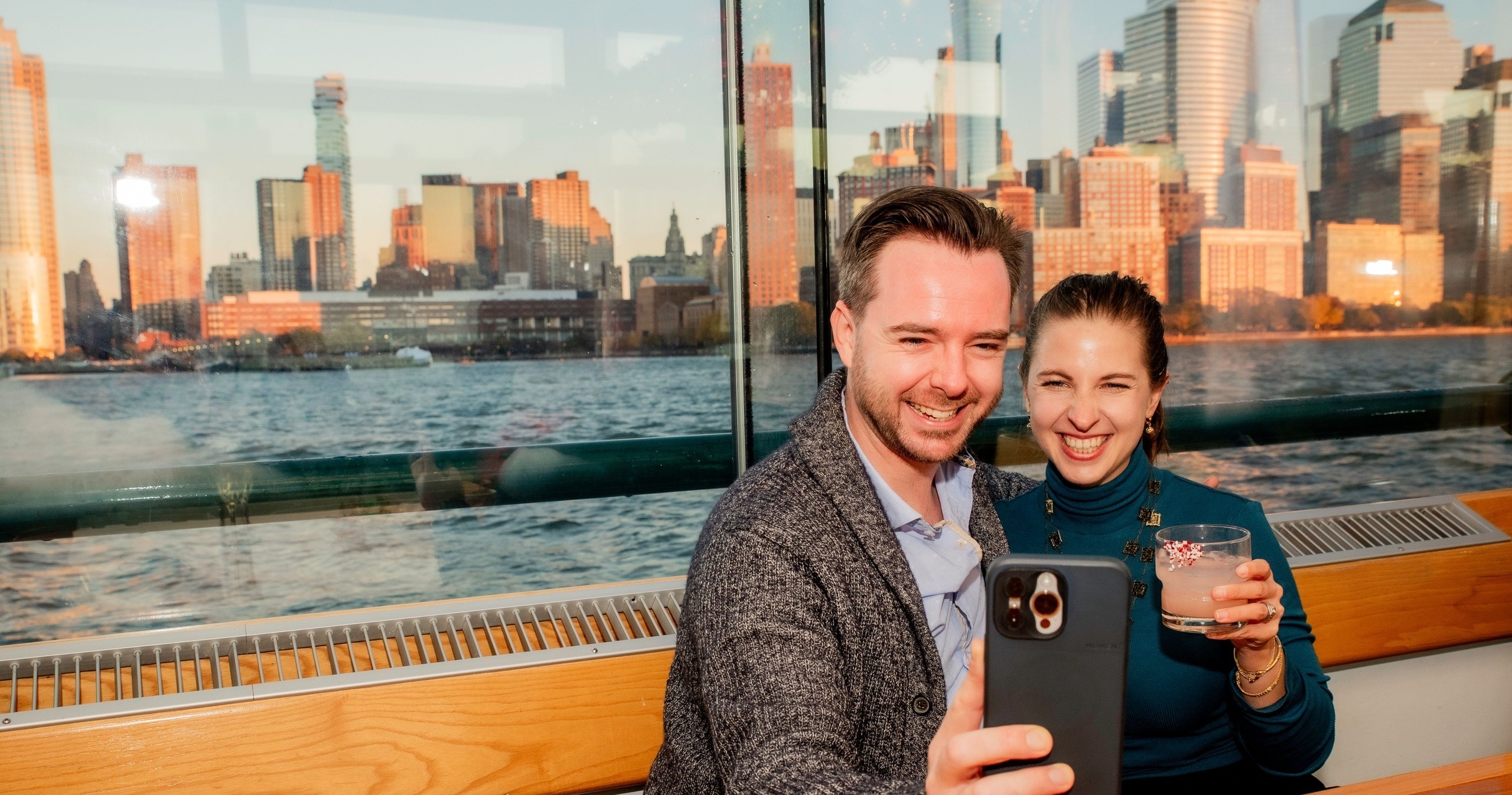 Couple taking a selfie on a boat with city skyline at sunset in the background.