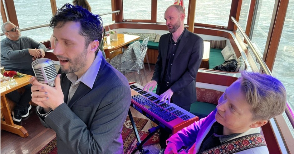 Three musicians performing on a boat, man singing, another playing keyboard, and third with a guitar.