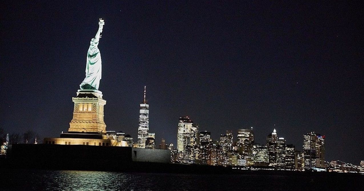 Illuminated Statue of Liberty at night with city skyline in background.