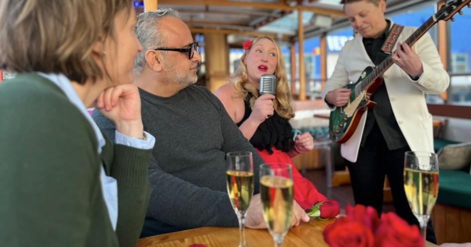 Woman singing with a microphone, guitarist playing, people listening, champagne glasses on table.