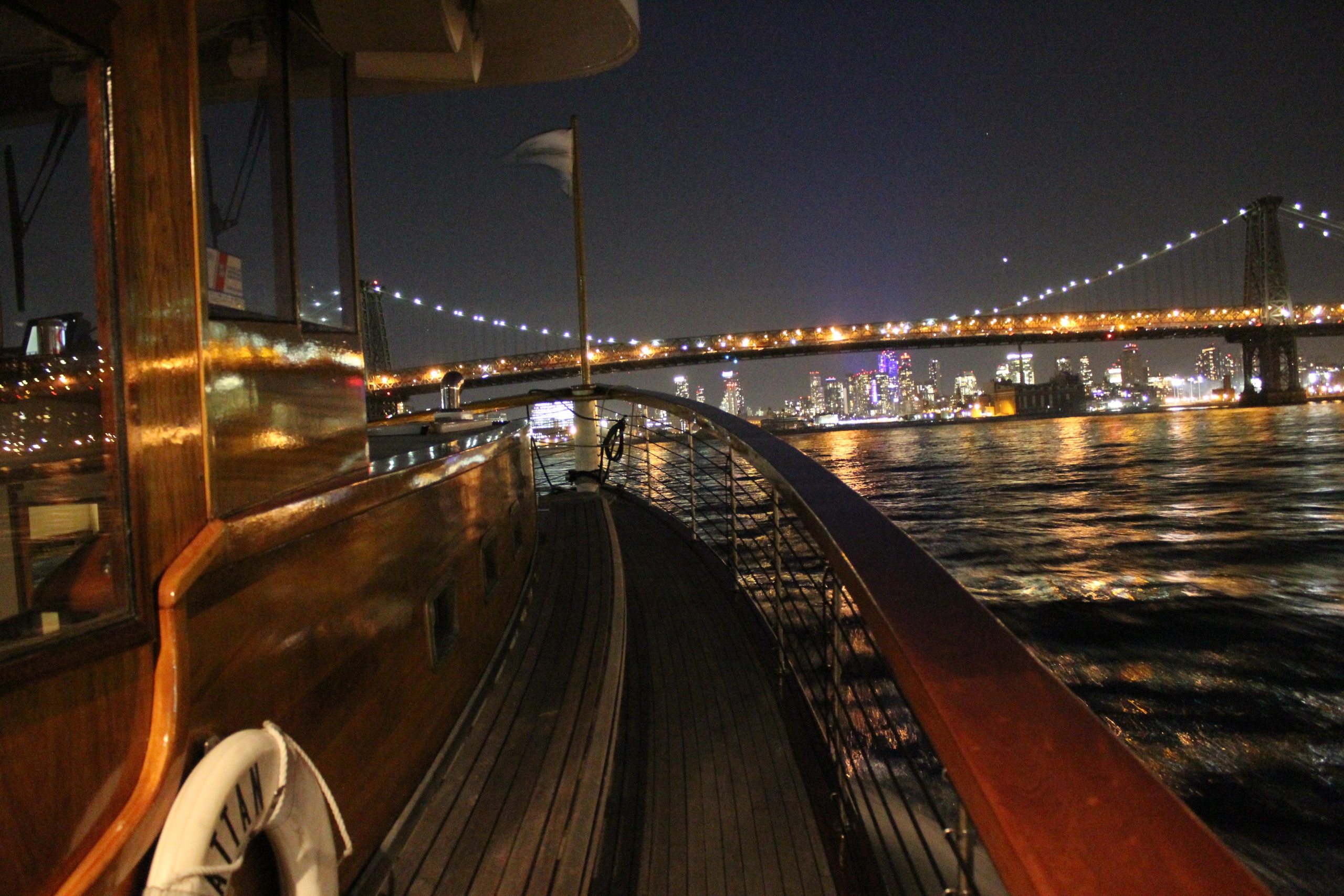 View from a boat deck at night with a city skyline and bridge in the background.