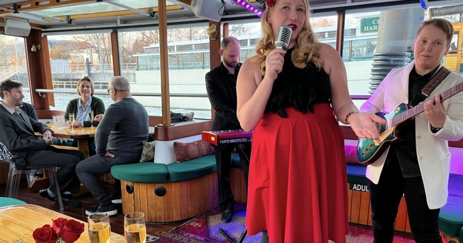Woman singing with a microphone on a boat, guitarist beside her; people seated at tables in the background.