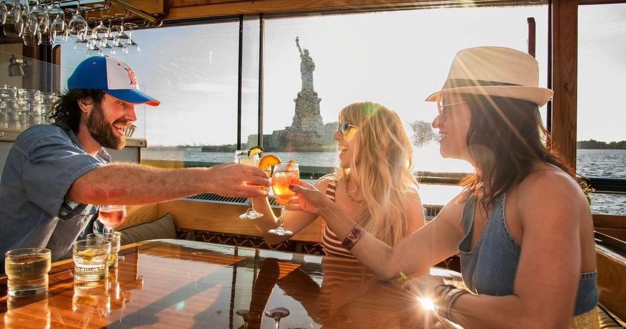 Three people on a boat toasting drinks with the Statue of Liberty in the background.