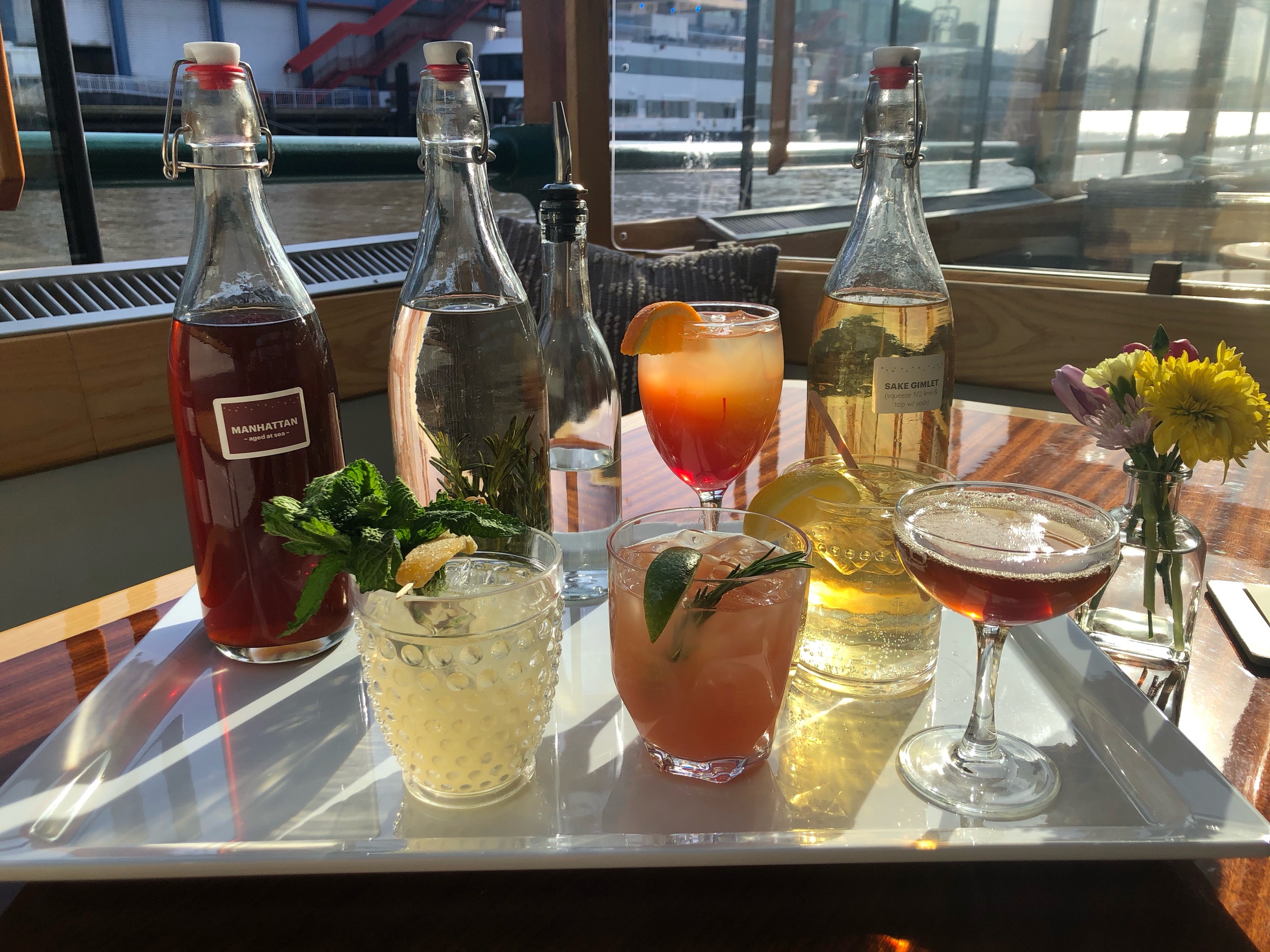 Assorted cocktails and bottles on a tray with flowers in sunlight.