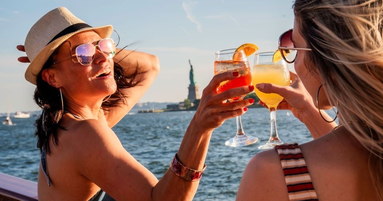 Two women toasting with drinks on a boat near the Statue of Liberty.