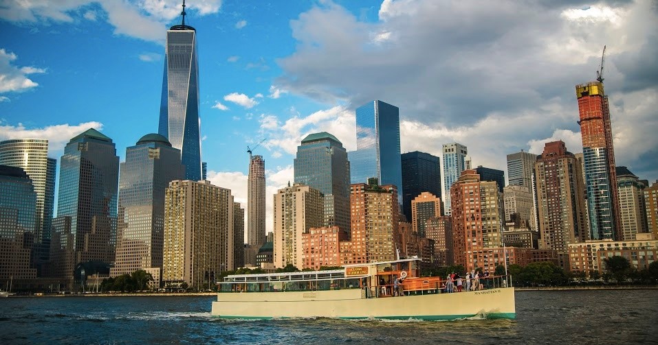 Boat sailing on river with city skyline in the background under a cloudy sky.