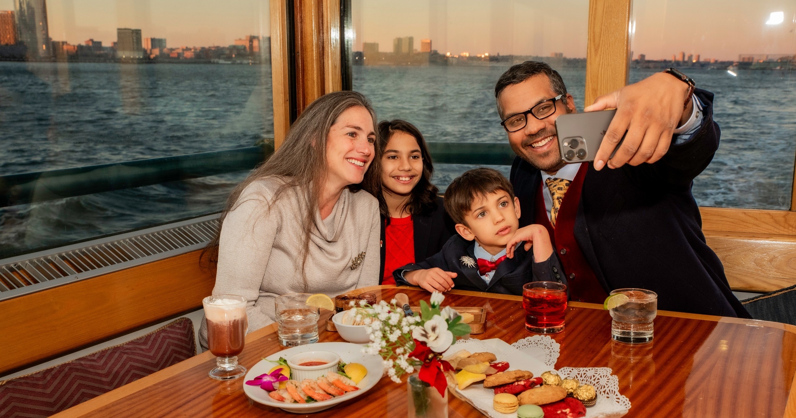 Family taking a selfie at a dining table on a boat with a city view.