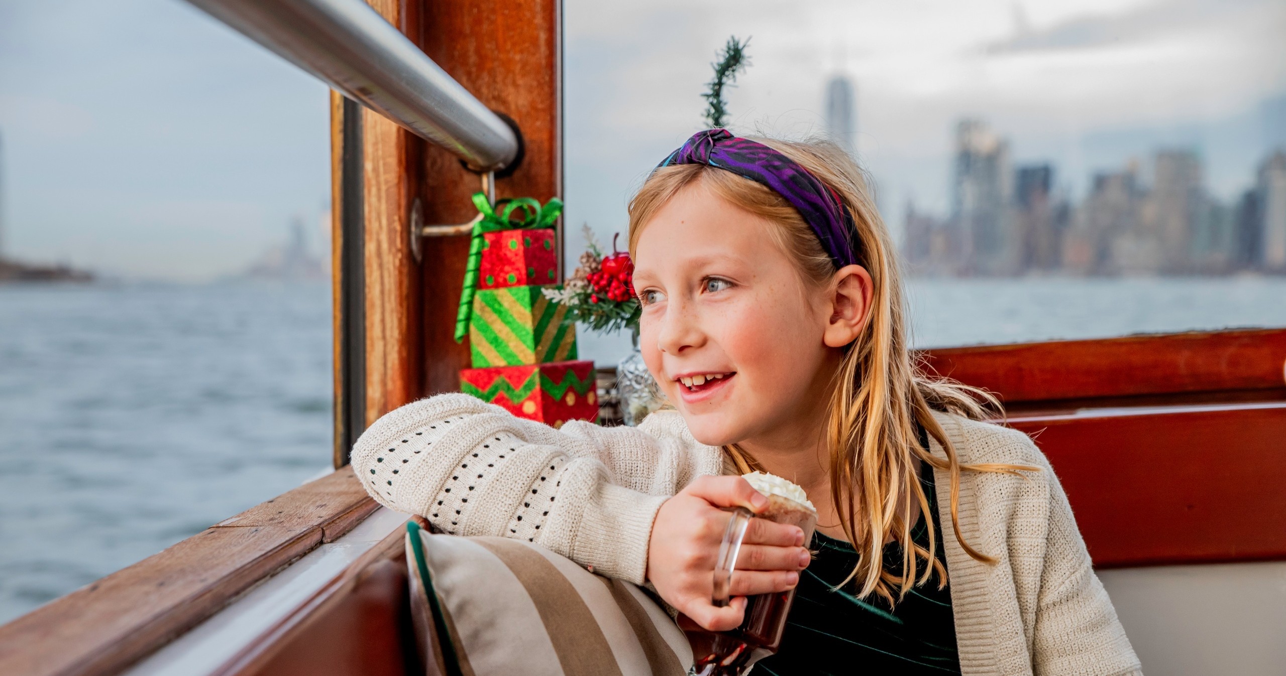 Girl on a boat with hot cocoa, city skyline in background.