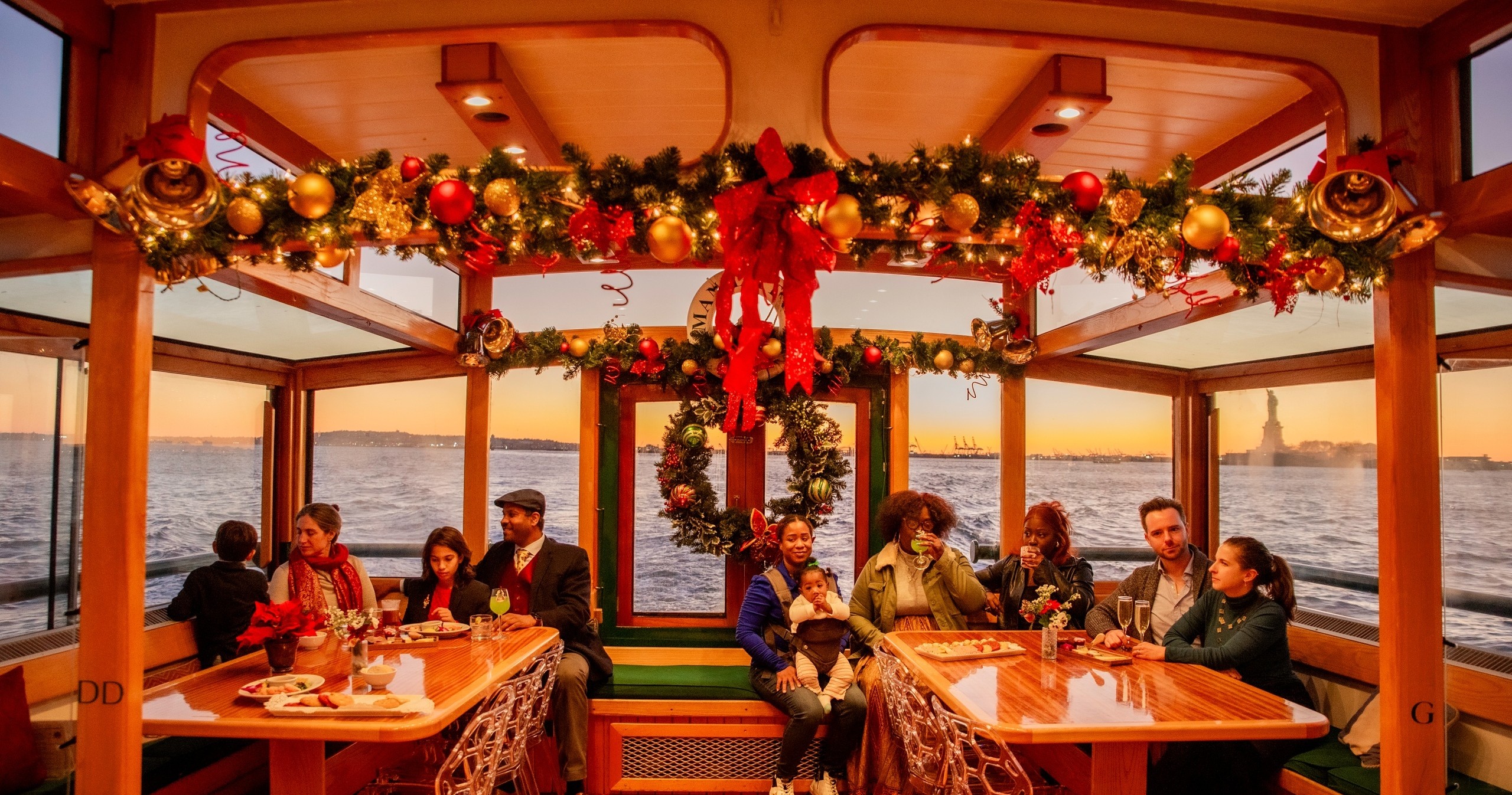 Festive boat interior with holiday decorations, people seated at tables, sunset view of water and distant statue.