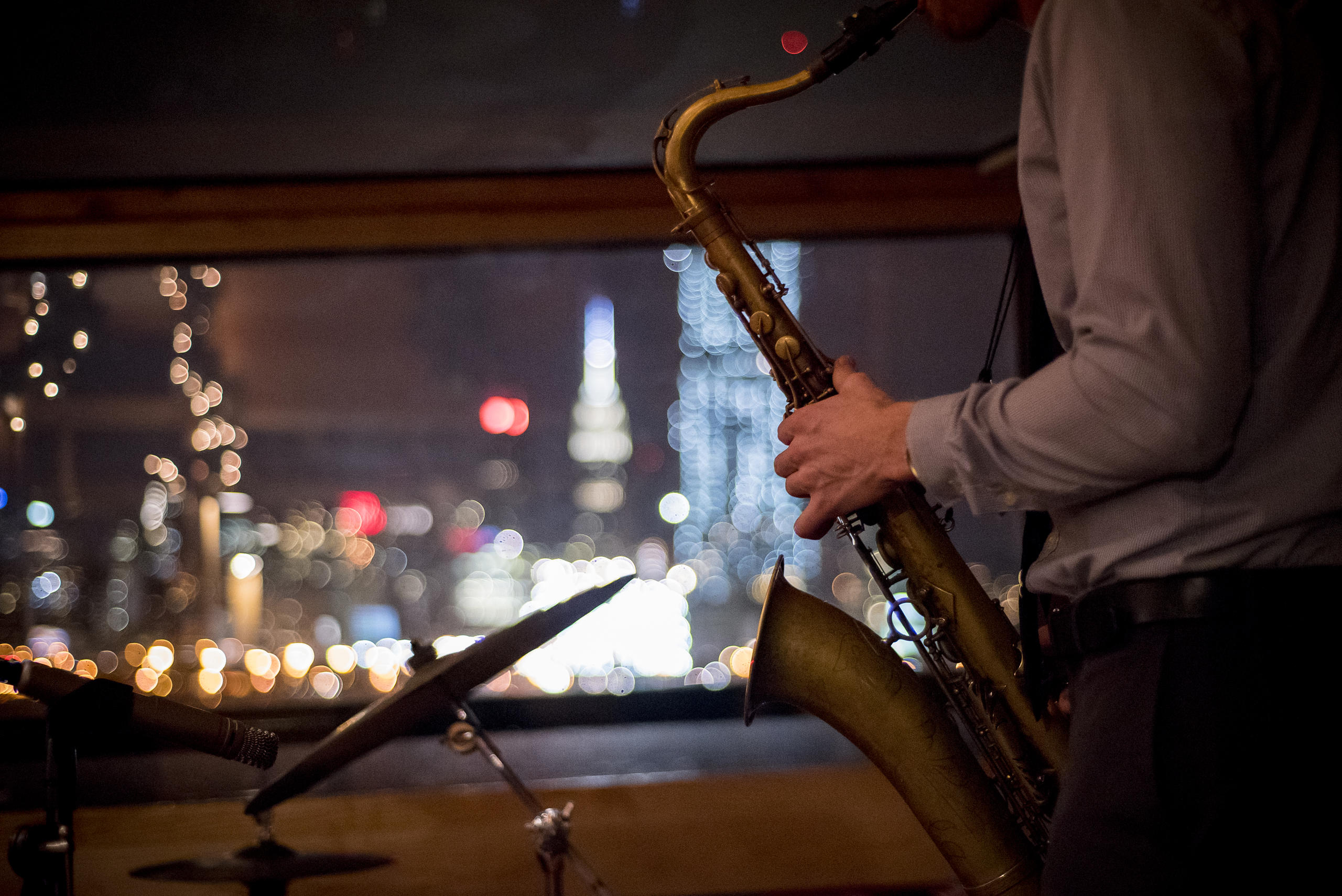 Man playing saxophone in dimly lit room with city lights outside window.
