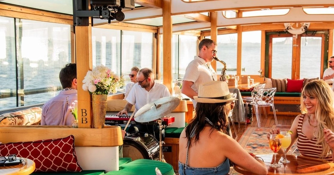 People enjoy music on a boat with a saxophonist and a drummer, surrounded by ocean views.