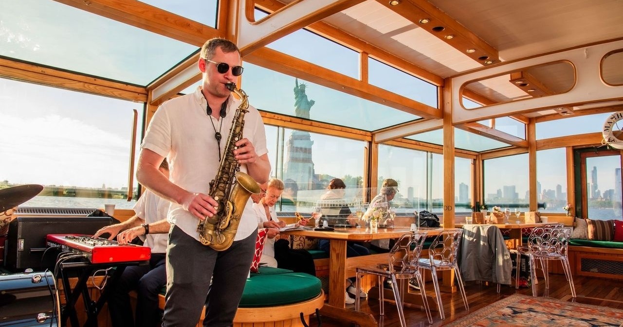 Man playing saxophone on a boat with the Statue of Liberty in the background.