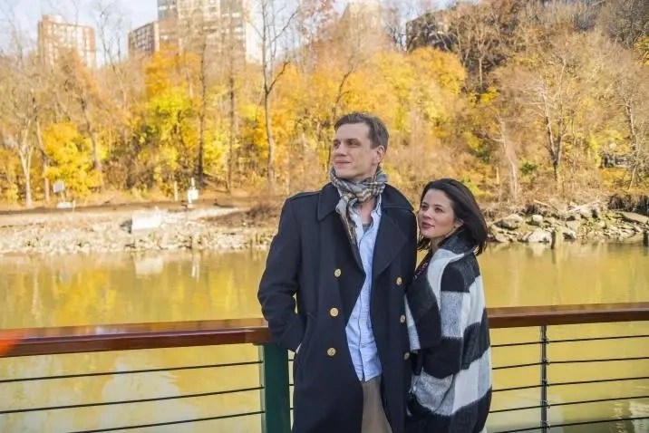 A couple stands by a railing with autumn trees and buildings in the background.