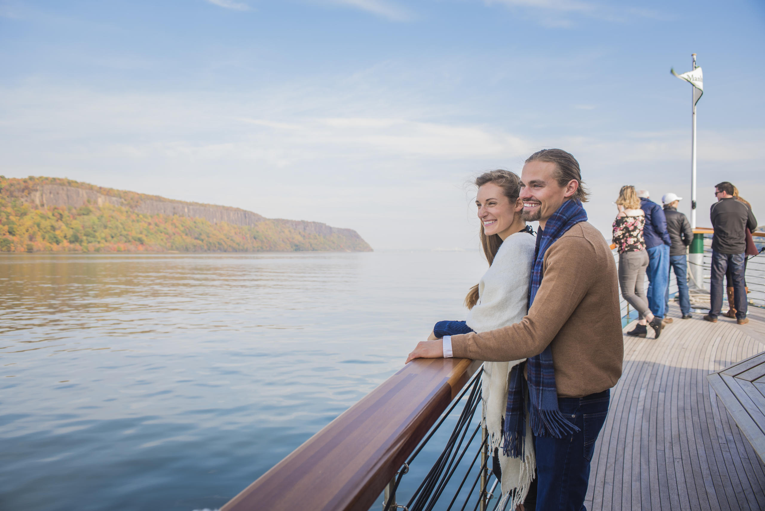 Couple on a boat deck enjoying a scenic view of the water and riverbank with others in the background.