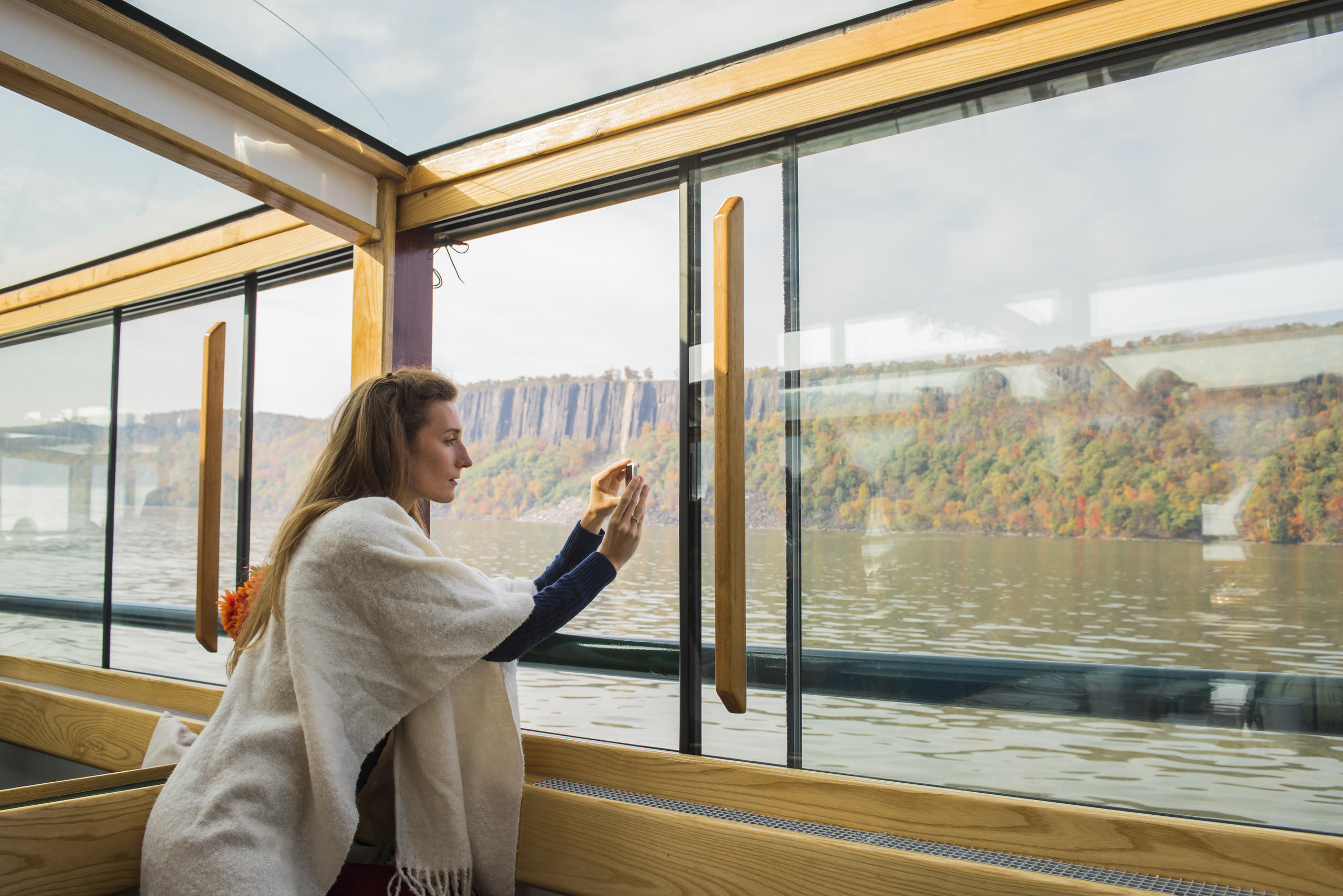 Woman in shawl taking photos on a boat with autumn trees and cliffs in the background.