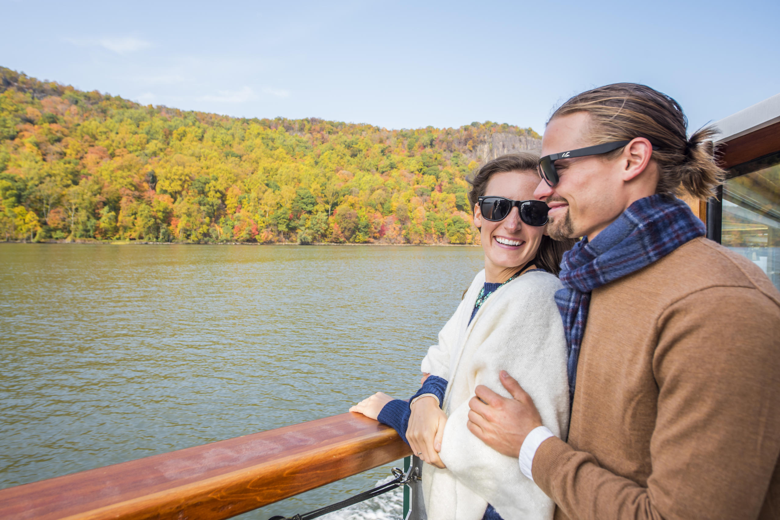 Couple in sunglasses on a boat, enjoying scenic lake and autumn trees.