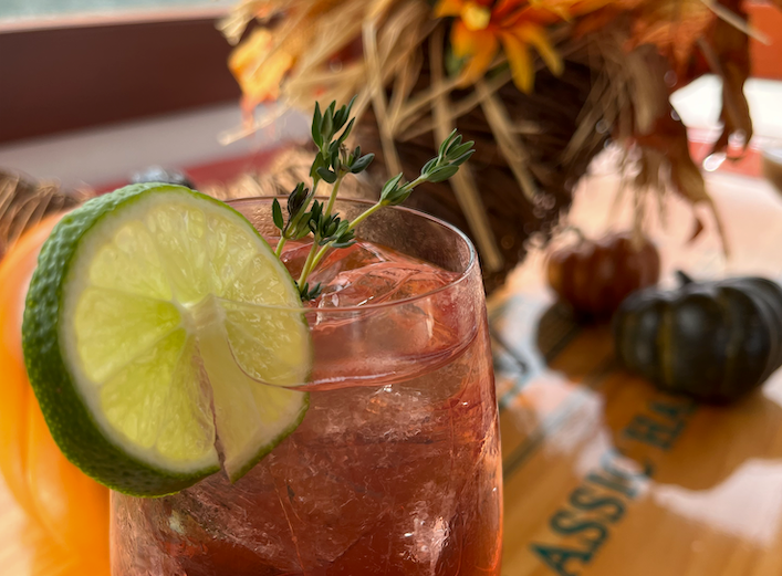 Cocktail with lime and herbs on table, fall decor in background.