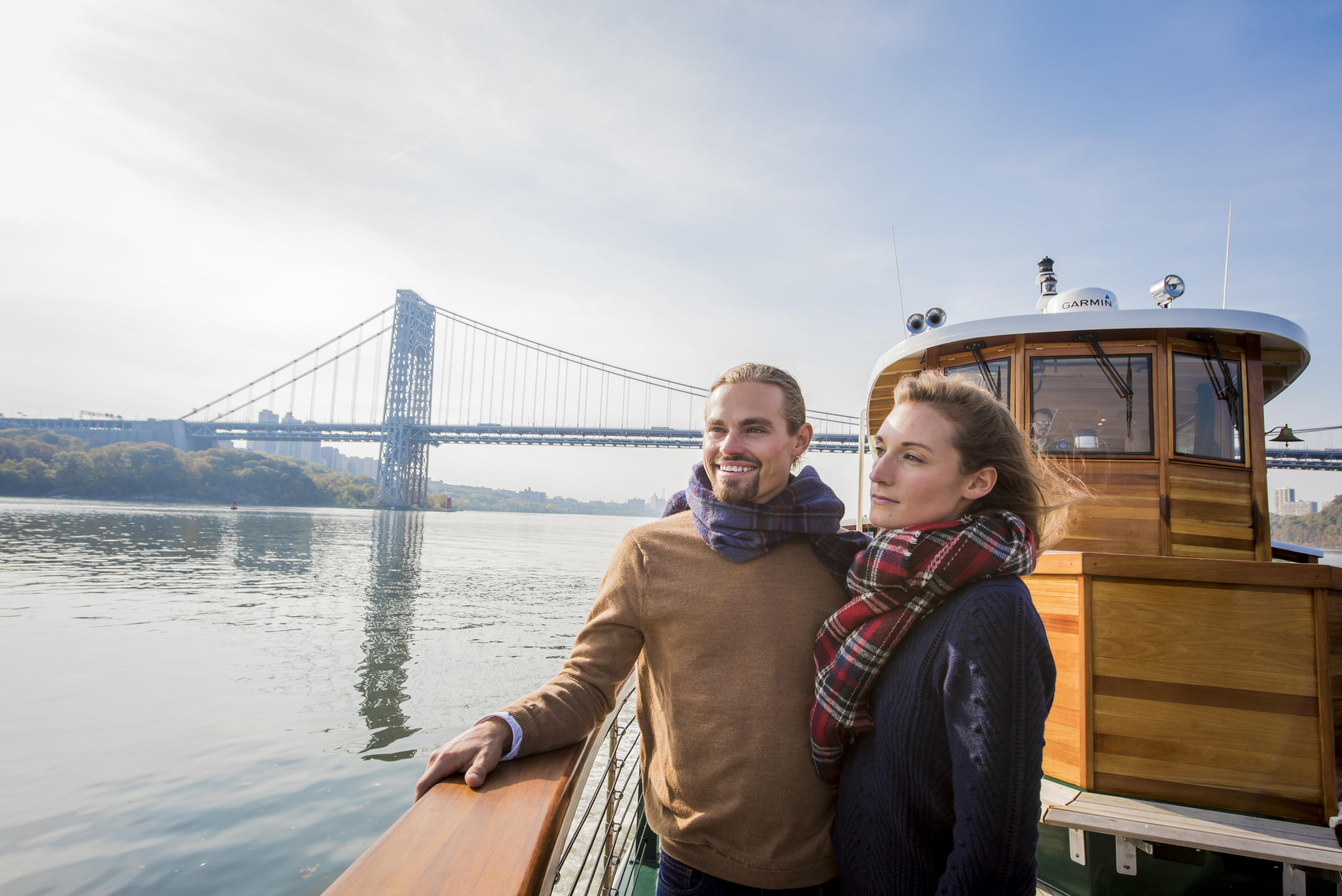 Couple on boat deck, river view, large bridge in background.