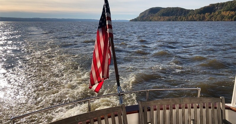 Boat with American flag on lake, surrounded by hills and a cloudy sky.
