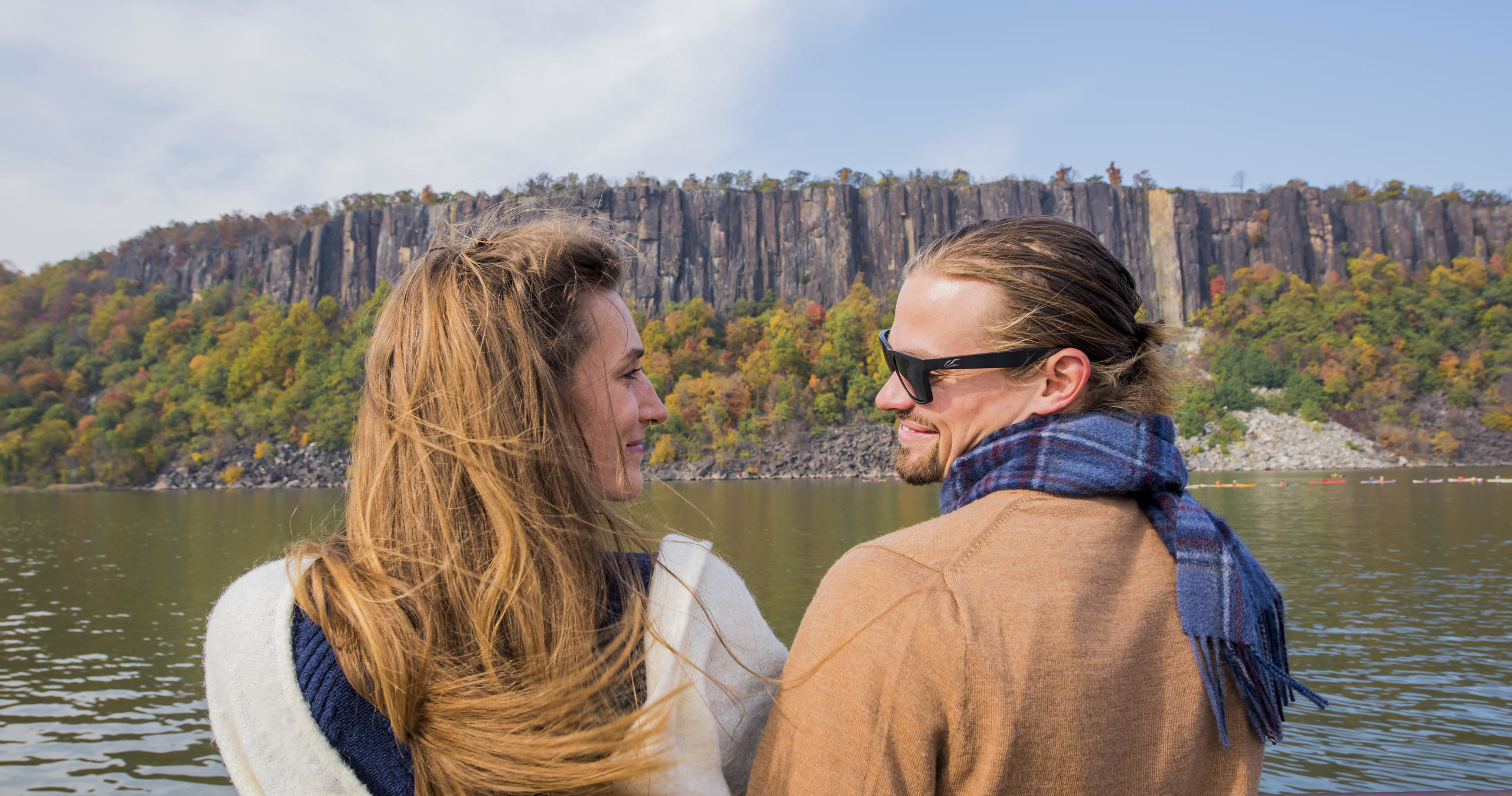 A couple looks at each other by a lake with a rocky cliff and autumn trees in the background.