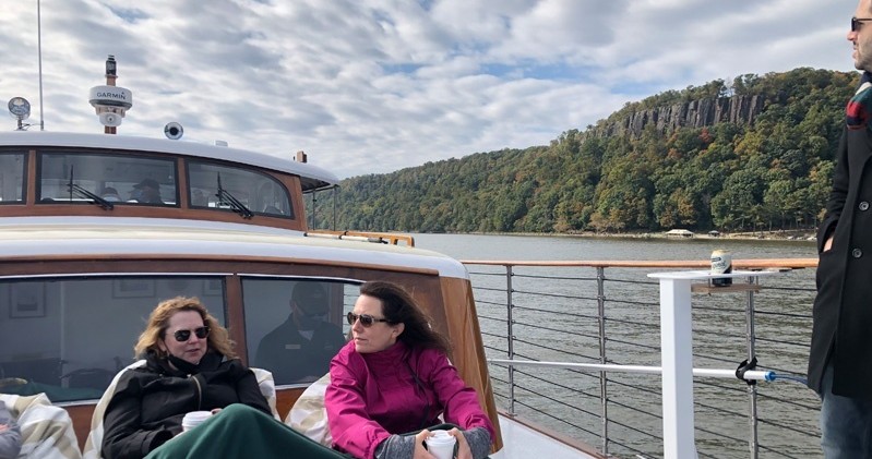 People relax on a boat with scenic river and forested cliffs in the background.