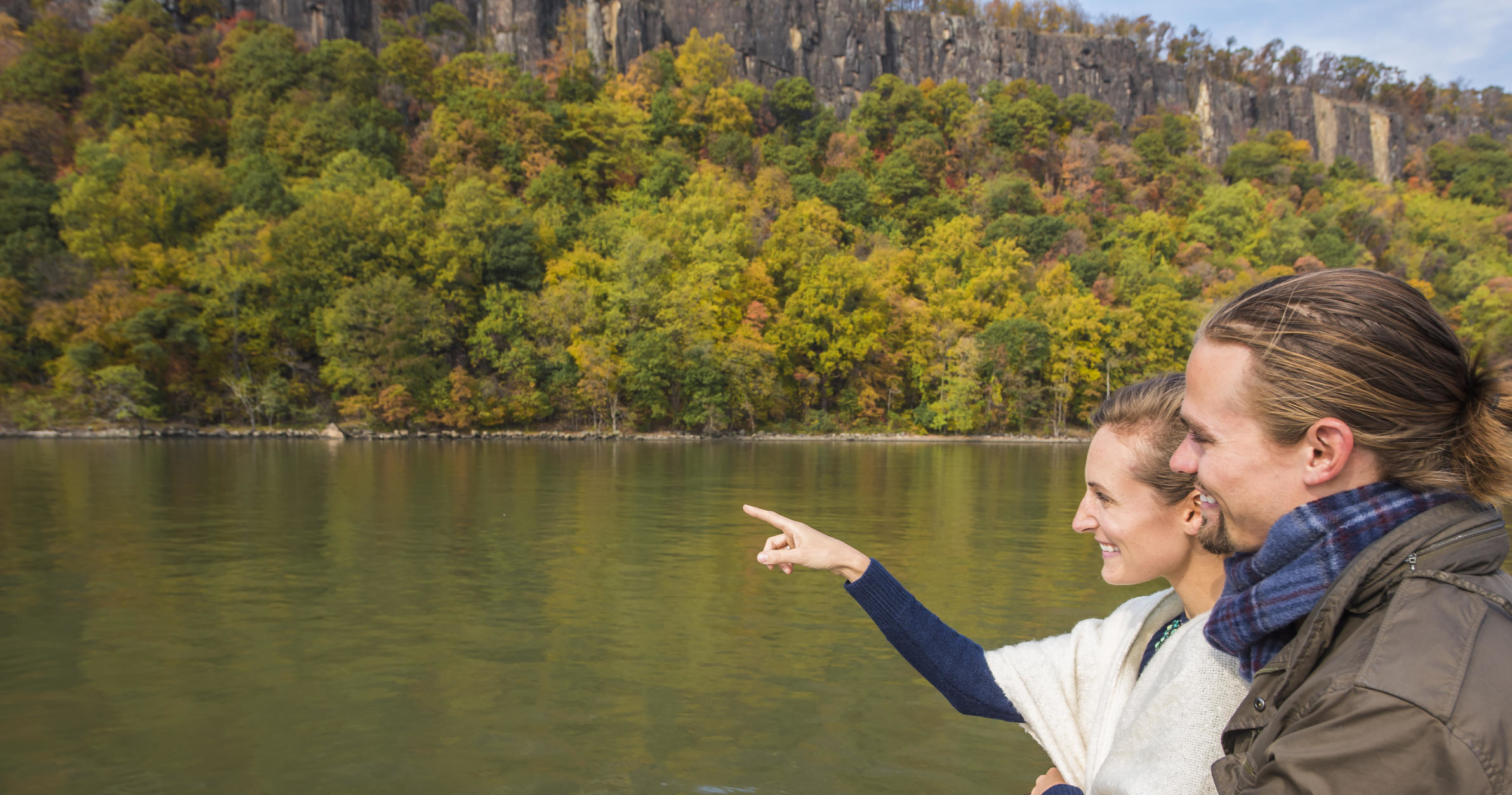 Two people on a boat, one pointing at a forested riverbank with cliffs.