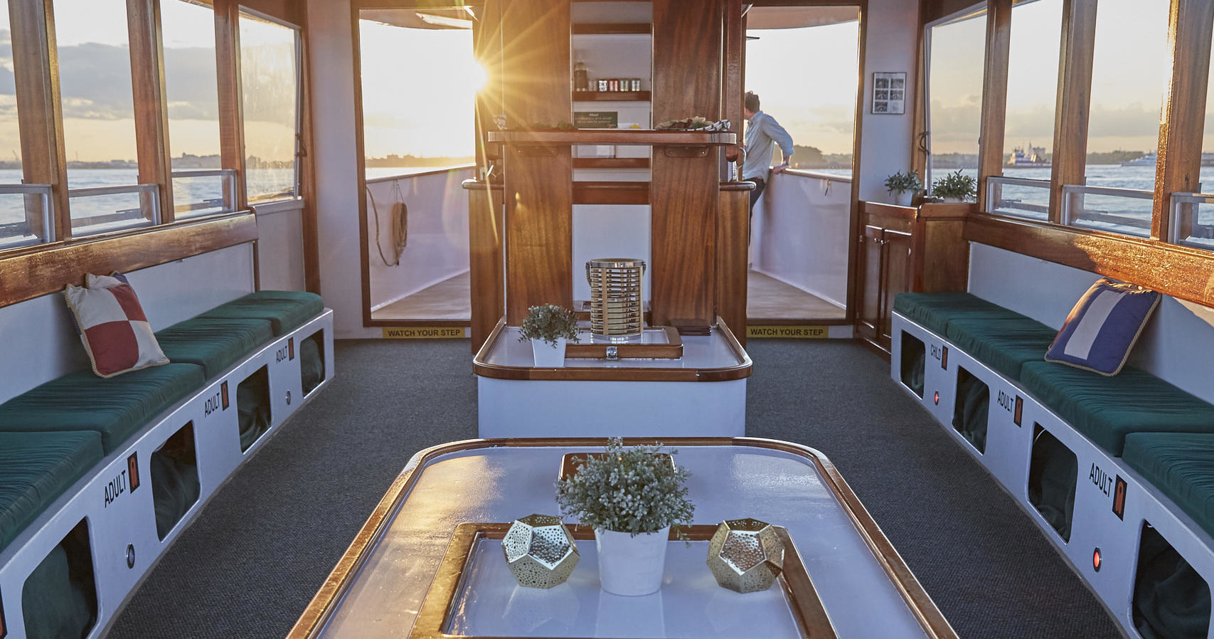 Sunlit boat interior with plants and seating, windows overlooking water.